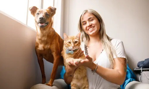 Happy woman holding her ginger cat and standing next to her dog, showcasing the joy of pet ownership and the value of hiring pet sitters, as discussed in our Ultimate Guide to Pet Sitting: Ensuring Your Pets Are Happy While You're Away.